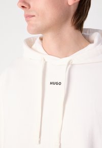 Jeune personne portant un sweat à capuche blanc avec le logo « HUGO » centré sous l'encolure, regardant vers la gauche sur un fond uni.