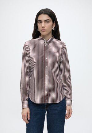 Jeune femme portant une chemise à rayures verticales marron et blanches et un jean bleu, debout devant un fond clair uni.