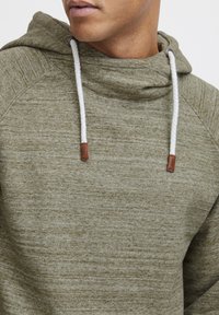 Sudadera con capucha verde oliva hecha de tela texturizada, con cordones blancos y acentos de cuero marrón, y un ajuste relajado.