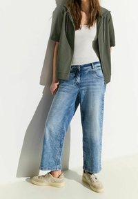 Oversized, light blue denim jeans with wide legs and faded texture, paired with a short olive green hooded jacket and beige sneakers.