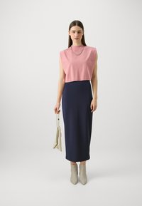 Rosa kort topp med korte ermer, kombinert med en tettsittende marineblå maxi-skjørt. Modellen holder en beige veske og har på seg lysegrå ankelstøvler.