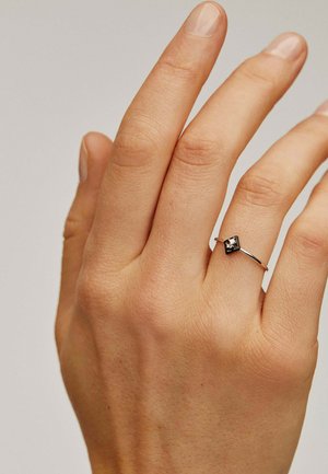 Anillo de plata con una piedra negra cuadrada, llevado en una mano. Banda delgada con un acabado suave y diseño minimalista. Textura de la piel natural visible.