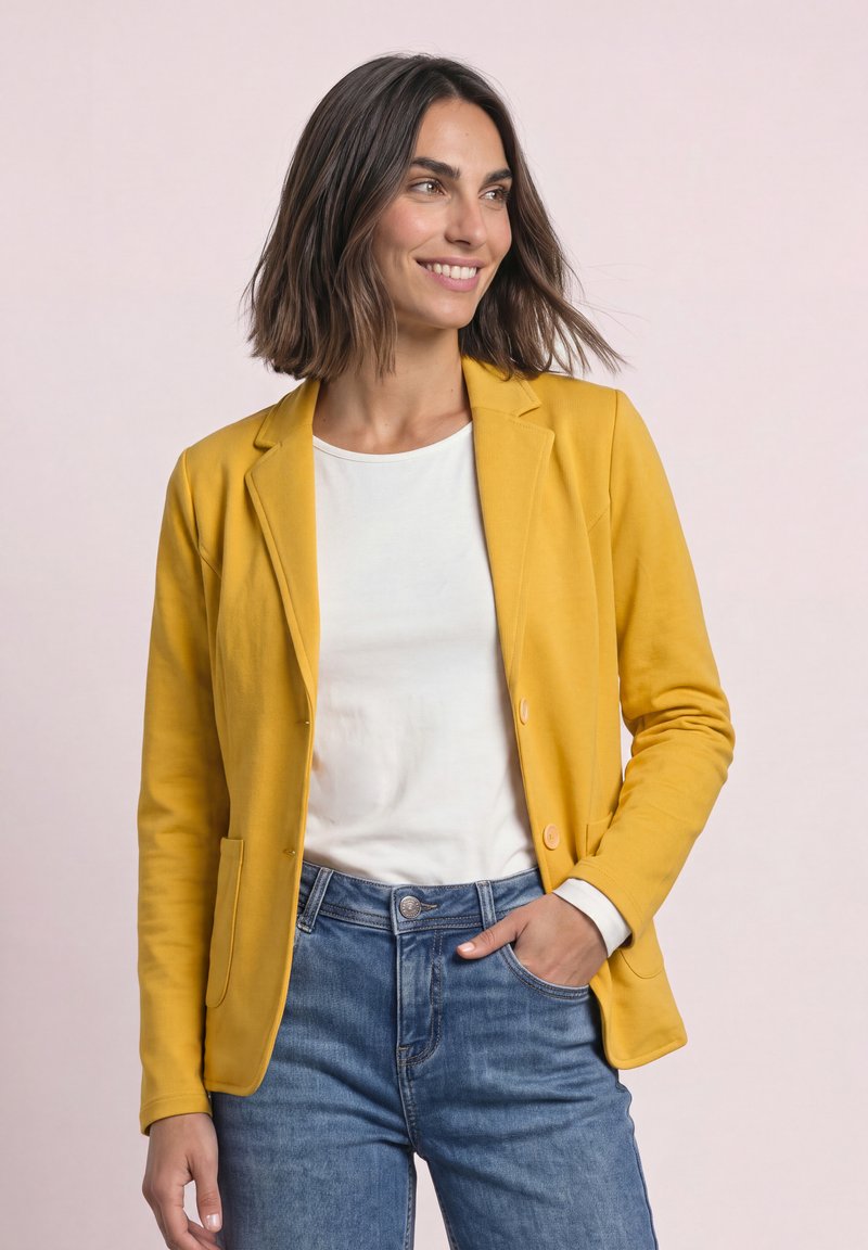 Femme souriante de profil portant un blazer jaune sur une chemise blanche et un jean bleu, avec une main dans la poche, devant un fond uni.