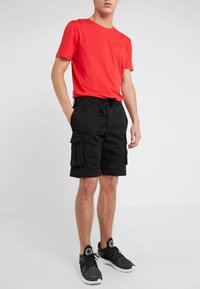 Červené bavlněné tričko s kulatým výstřihem, spárované s černými cargo šortkami s bočními kapsami. Černé tenisky dokončují celý outfit.