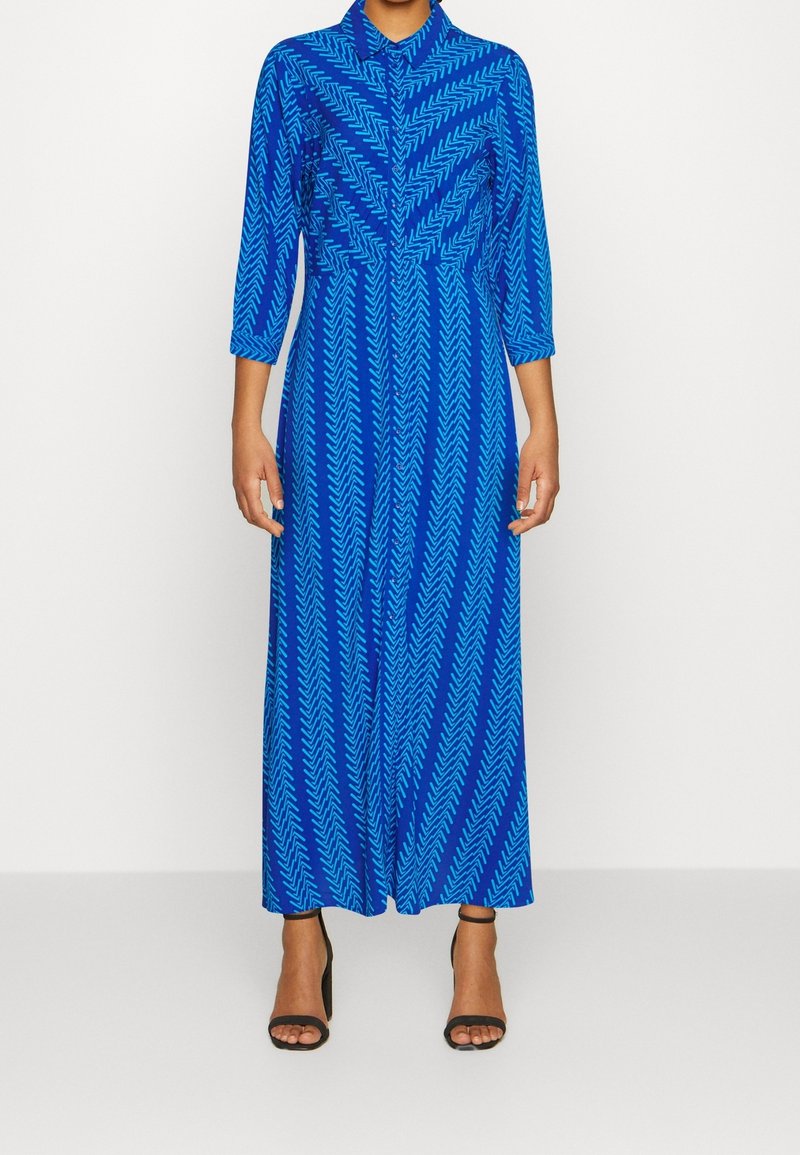 Femme portant une longue robe bleue avec des motifs chevrons bleu clair et des manches trois-quarts, associée à des talons noirs à bout ouvert.