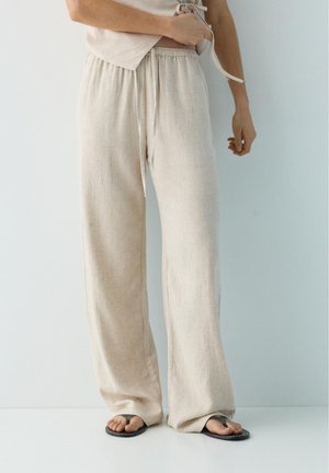 Široke bež lanene hlače s elastičnim pojasom i vezicom, nošene s crnim japankama na svijetloj pozadini.