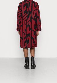 Roter und schwarzer Mantel mit geometrischem Muster, langen Ärmeln und lockerem Schnitt, kombiniert mit schwarzen Stiefeletten, Ansicht von hinten.