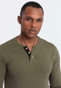 Mann mit kurzem Haar, der ein olivgrünes Langarm-Henley-Shirt mit drei Holzknöpfen und einer schwarzen Knopfleiste trägt.