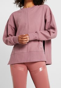 Osoba v broskvově růžovém oversize svetru a odpovídajících růžových legínách, ruce složené v pase, kudrnaté vlasy viditelné nad límcem.