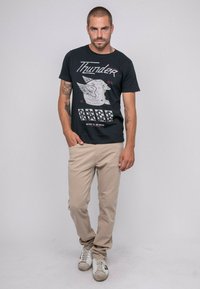 T-shirt noir avec un design graphique comprenant des ailes et du texte, associé à un pantalon beige et des baskets blanches avec des accents noirs.