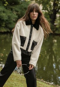 Veste en faux fur et cuir, blanche et noire, avec de grands boutons, deux poches avant et un col. Modèle tenant des lunettes de soleil blanches.