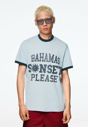 Homme aux cheveux courts décolorés, portant des lunettes de soleil bleues et un t-shirt bleu clair avec le texte "Bahamas Sunset Please" et un short rouge.