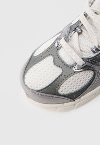 Ελαφρύ αθλητικό παπούτσι με άνω μέρος από λευκό δίχτυ, γκρι συνθετικές λεπτομέρειες, υφή επιφάνειας και επίπεδα κορδόνια.