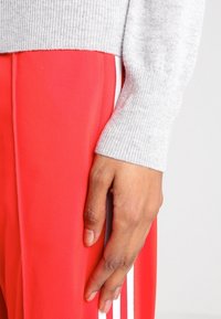 Manche de pull rigide gris aux côtés de pantalons de sport rouge vif avec des rayures blanches; main visible reposant sur la jambe du pantalon.