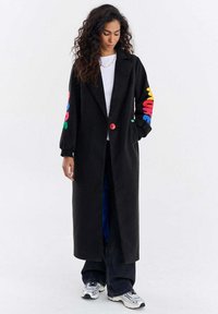 Kobieta o długich kręconych włosach, ubrana w czarny długi płaszcz z kolorowymi literami na rękawach, białą koszulę, ciemne spodnie i białe trampki, stoi.