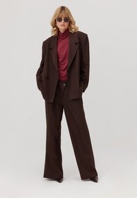 Kobieta o blond włosach, ubrana w dużą ciemnoniebieską marynarkę, bordowy golf, okulary przeciwsłoneczne i złote kolczyki, stoi z rękami w kieszeniach.