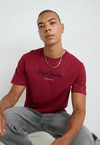 Κόκκινο κοντομάνικο μπλουζάκι με λογότυπο "Pepe Jeans London" σε μαύρο χρώμα. Το μοντέλο φοράει γκρι τζιν. Λείο ύφασμα και κλασικός σχεδιασμός με στρογγυλή λαιμόκοψη.