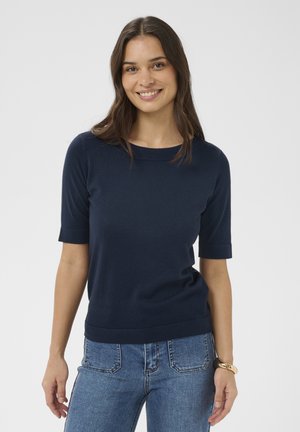 Mujer con cabello largo y castaño sonriendo, vistiendo una camiseta de manga corta azul marino y jeans azul claro, con una pulsera negra y dorada en la muñeca.