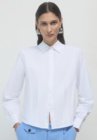 Chemise blanche à manches longues avec col classique, pr�ésentant des détails plissés et un ourlet légèrement court, portée avec un pantalon bleu.