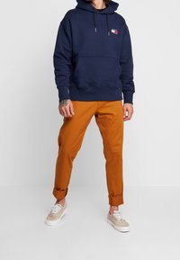 Sudadera con capucha azul marino con bolsillo delantero y cordón, combinada con pantalones naranjas con puños enrollados y zapatillas beige con cordones blancos.