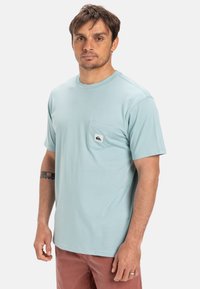 Světlomodré tričko s krátkými rukávy z měkké látky, s přední náprsní kapsou a bílou a černou logo nášivkou.