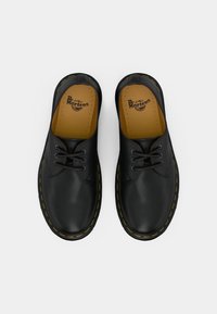 Pár černých kožených bot Dr. Martens s žlutým švem a černými tkaničkami, zobrazených z výšky na bílém pozadí.