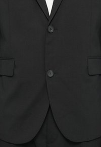 Chaqueta de traje negra hecha de tela suave, con dos botones frontales, dos bolsillos y un sutil patrón de rayas en el material.