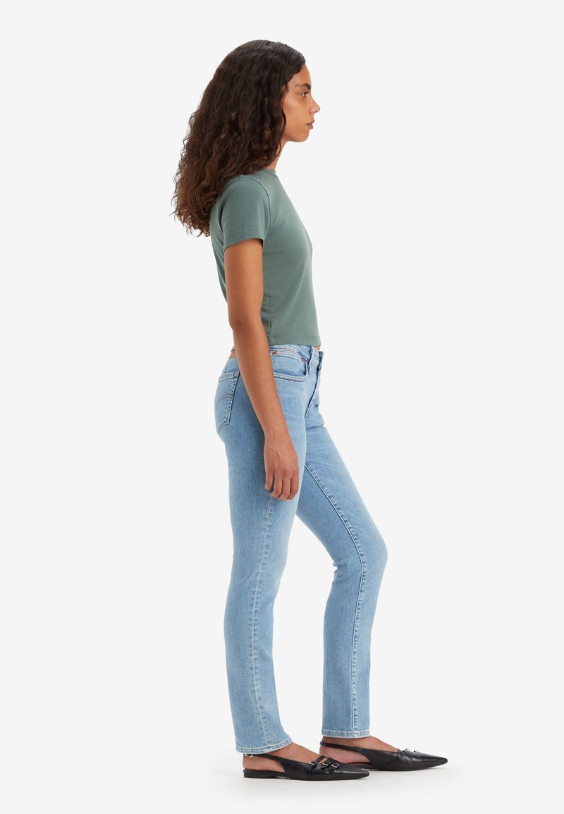 Camiseta verde ajustada, jeans skinny azul claro y zapatos negros sin cordones. Modelo mostrado de lado contra un fondo blanco.
