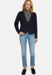 Blazer bleu marine à coupe ajustée, fermeture à un bouton et revers crantés, associé à un col roulé rayé et un jean bleu clair retroussé.