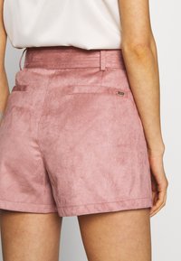 Růžové semišové šortky s hladkou texturou, vysokým pasem a dvěma zadními kapsami. Obsahují malou značkovou visačku na zadní straně.