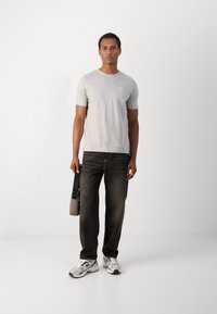 Tricou gri cu mânecă scurtă, blugi închiși la culoare și adidași gri. Persoana ține o geantă mică de umăr, bej. Îmbrăcăminte casual cu o croială relaxată.
