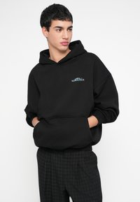 Hanorac negru din material moale, cu buzunar frontal și logo brodat "WORLD TOUR" în albastru. Croială lejeră, cu glugă spațioasă.