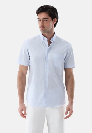 Mann trägt hellblaues kurzärmeliges Hemd mit Brusttasche und weiße Hose, steht vor einem einfarbigen weißen Hintergrund.