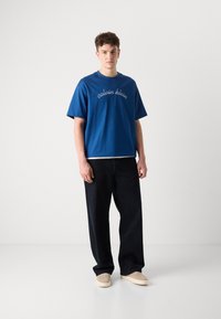 Mėlyna medvilninė marškinėliai su balta "calvin klein" siuvinėjimu, derinami su laisvai krentančiomis tamsiomis džinsomis ir beige slip-on bateliais. Sklandi tekstūra.