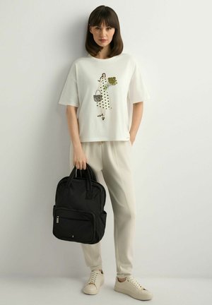 Kobieta stojąca przy ścianie, ubrana w białą koszulkę z grafiką kobiety, beżowe spodnie, białe trampki, trzyma czarny plecak za uchwyty.