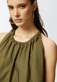 Maslinasto zelena bezrukavna majica s nabranim izrezom i mekim teksturom, koja ima opušteni kroj koji se nježno drapira preko tijela.