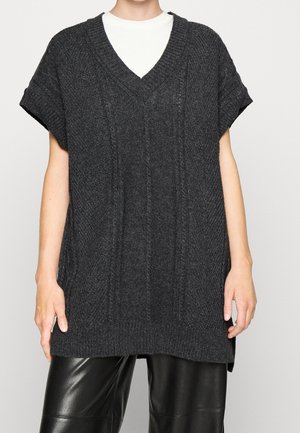 Gilet en tricot gris foncé à manches courtes et col en V porté sur une chemise blanche avec un pantalon en cuir noir, sur une personne debout devant un fond clair.