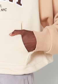 Felpa crema e beige realizzata in morbido tessuto, con tasca frontale e polsini a coste, caratterizzata da una scritta ricamata in contrasto sul petto.