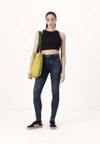 Black cropped tank top, blue skinny jeans, black sneakers with white stripes, and a yellow pleated shoulder bag. Braided hair and necklace visible.