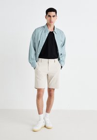 Stripete grønn og hvit langermet skjorte over en svart t-skjorte, sammen med lyse beige shorts og hvite sneakers med teksturert såle.