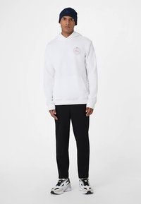 Hvid hættetrøje med rundt logo, kombineret med sorte taperede bukser og sorte og hvide sneakers. Beanie er marineblå og fuldender outfittet.