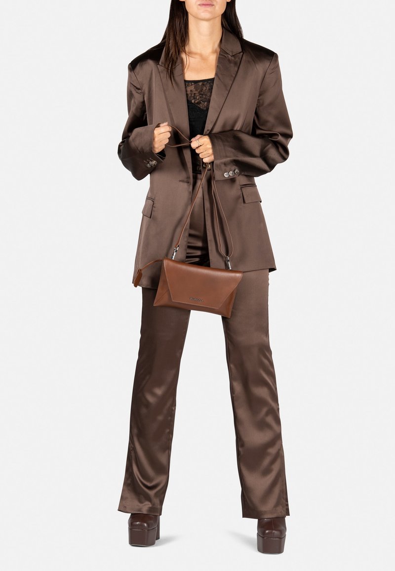 Femme en costume en satin marron avec un top en dentelle noire, tenant un sac bandoulière en cuir marron, debout sur des talons compensés.