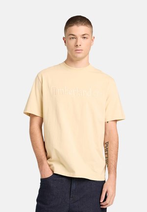 Giovane uomo con capelli corti che indossa una maglietta Timberland beige e jeans scuri, in piedi con una mano in tasca e espressione neutra.