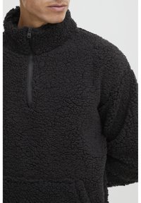 Sort fleece pullover med høj krave, med en halv lynlås foran, tekstureret stof og en kengurulomme. Blødt og hyggeligt materiale.