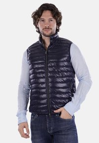 Gilet imbottito blu navy, design trapuntato, leggero, con zip frontale, colletto alto, abbinato a una camicia a maniche lunghe azzurro chiaro, indossato sopra dei jeans.