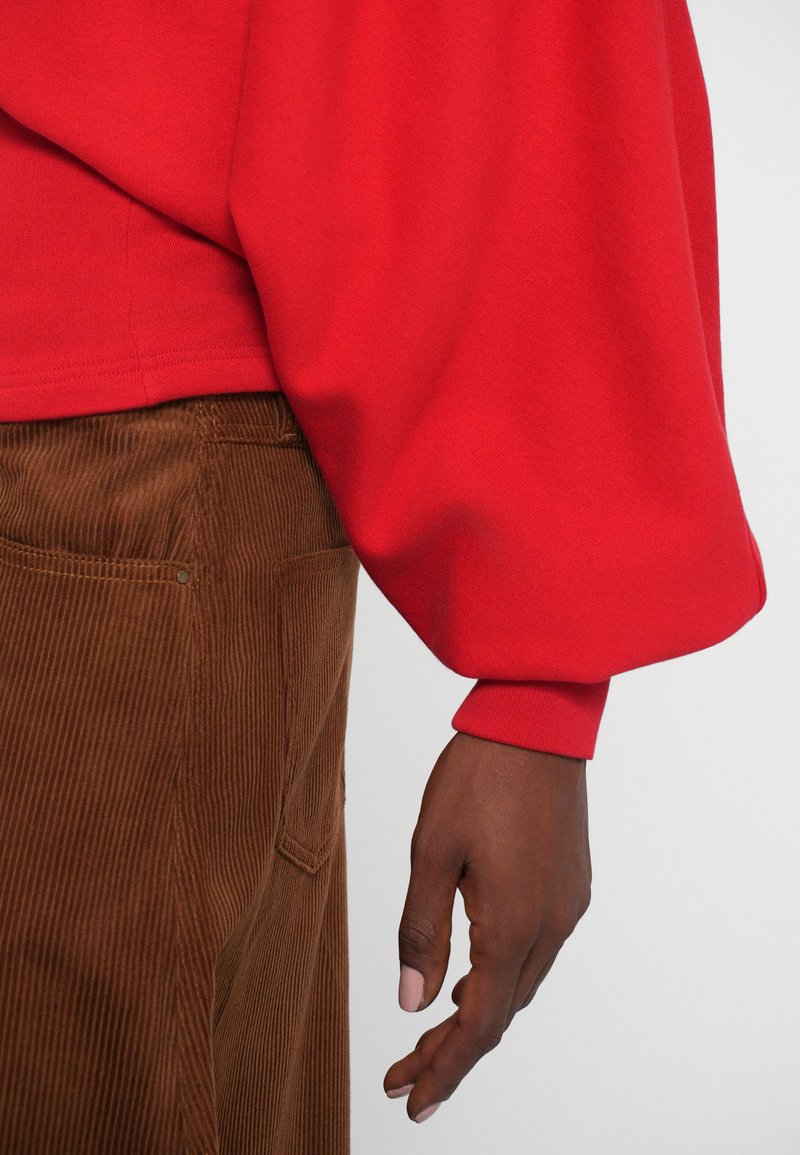 Sweatshirt rouge avec des manches ballon, fabriqué en tissu doux, associé à un pantalon en velours côtelé marron présentant un motif texturé à rayures verticales et des poches.