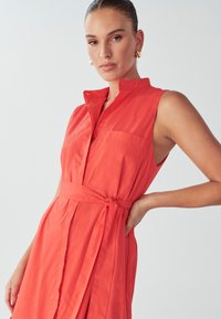 Koralen obleka s ovratnikom, gumbi spredaj, kratkimi rokavi in pasom. Ima stransko žep in gladko teksturo tkanine.