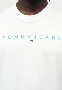 Λευκή βαμβακερή μπλούζα με το κέντημα "TOMMYJEANS" σε τιρκουάζ χρώμα και μια μικρή λεπίδα με μπλε, κόκκινο και λευκό λογότυπο κάτω από το κείμενο.