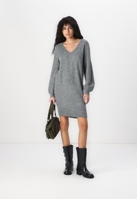 Robe tricotée grise avec un col en V, des manches longues et une coupe décontractée. Portée avec des bottes noires au genou et tenant un sac vert. Texture lisse.