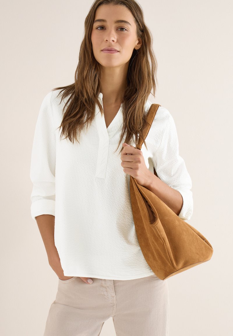 Jeune femme aux longs cheveux bruns portant un chemisier blanc texturé et un pantalon beige, tenant un grand sac à bandoulière en daim marron.